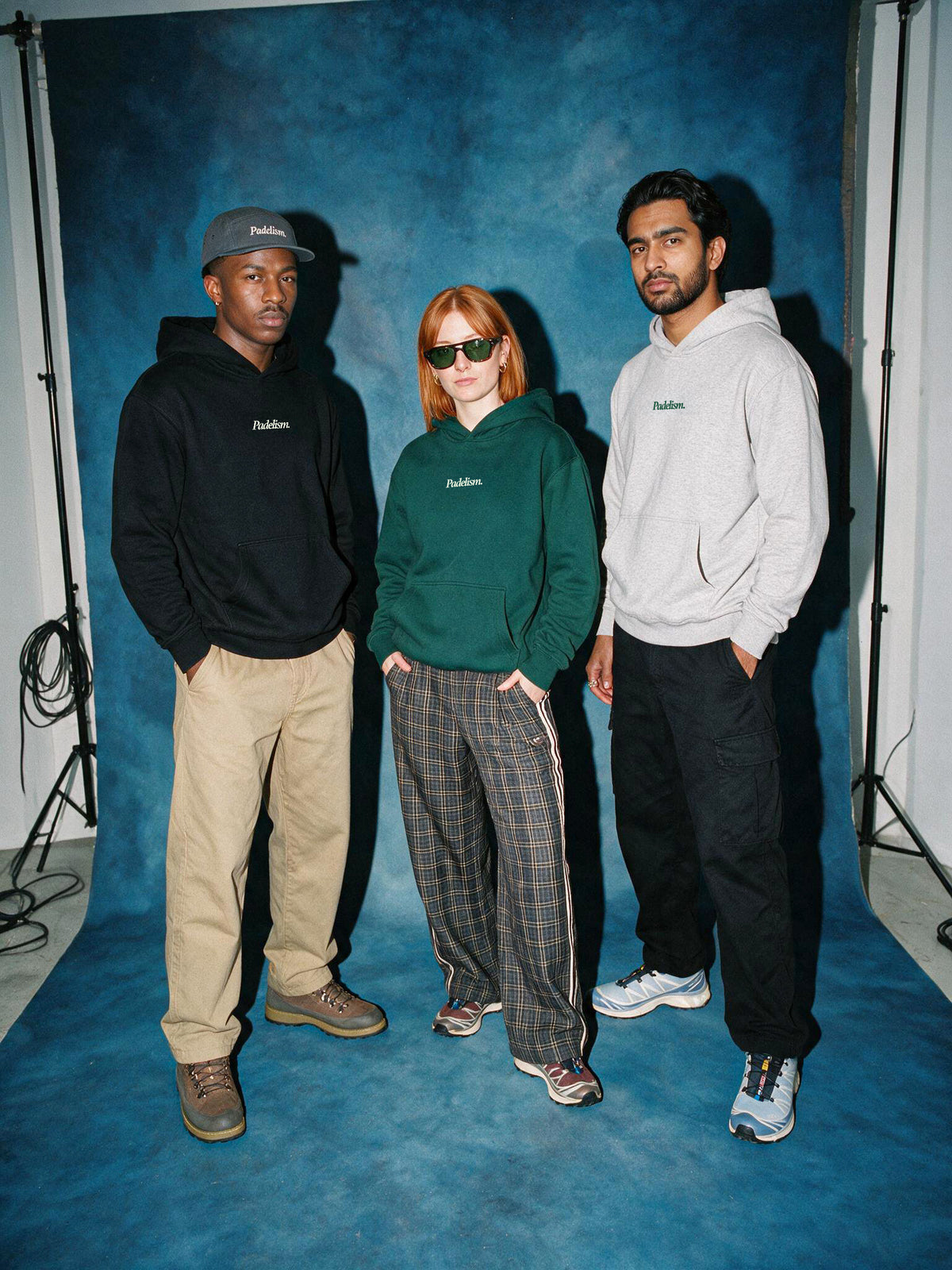 Three models in Padelism Mk1 Box Hoodies in Black, Eden Green and Flower Gray on a blue studio backdrop