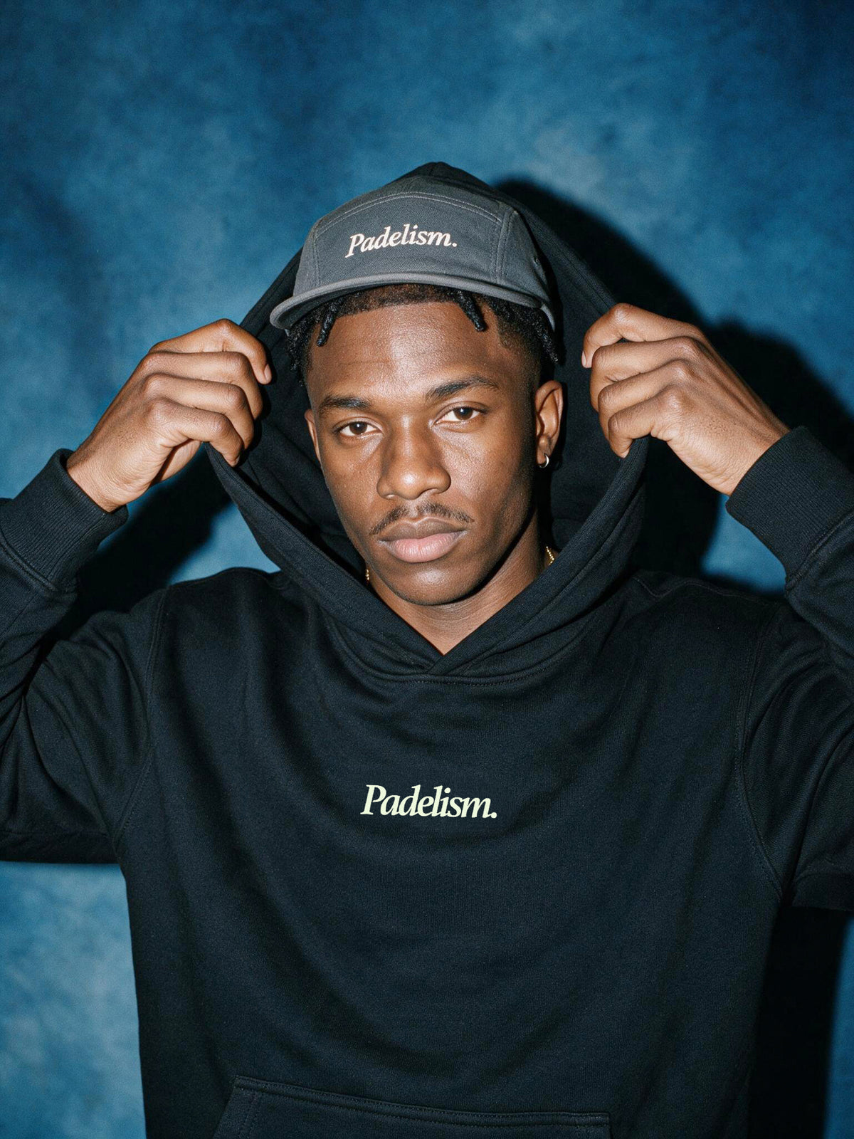 Model pulling up the hood of a black Padelism Mk1 Box Hoodie, wearing a Padelism cap